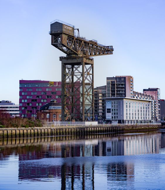 A tall, industrial crane structure painted in dark colors positioned next to a river, with several modern buildings in the background, including a distinctive purple and pink multi-story building and 