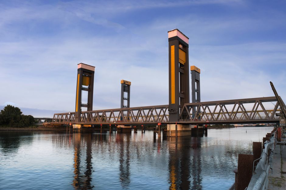 A tall, industrial crane structure painted in dark colors positioned next to a river, with several modern buildings in the background, including a distinctive purple and pink multi-story building and 